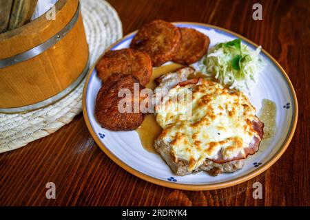 Pork steak with baked cheese and ham and small potato pancake on plate on wooden table with container with cutlery, rural. Stock Photo