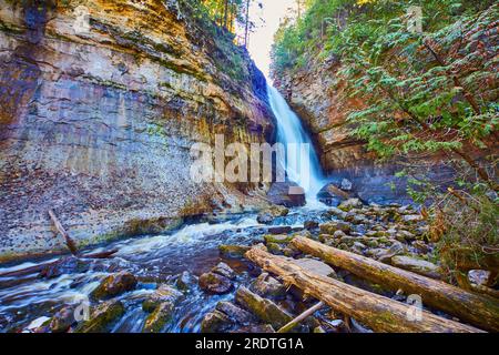 Waterfall over tall cliffs in canyon with greenery and driftwood in a dried-up riverbed over stones Stock Photo