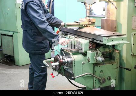 A male worker works on a larger metal iron locksmith lathe, equipment for repairs, metal work in a workshop at a metallurgical plant in a repair produ Stock Photo