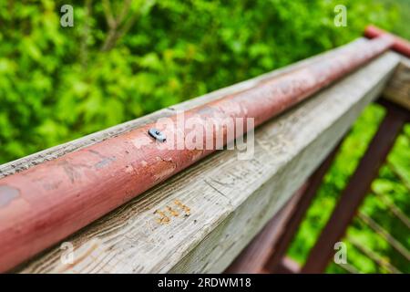 Chipped red paint on metal pole with lone screw atop wood railing with blurry green background Stock Photo