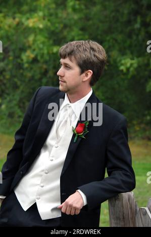 Groom, dressed in black tuxedo with with vest, shirt and tie, leans against a rustic wooden fence post.  He is solemn and thinking about his wedding d Stock Photo