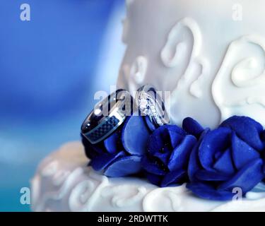 Two wedding rings nestle amoung blue roses on a wedding cake.  Cake is tiered and decorated with swirls. Stock Photo