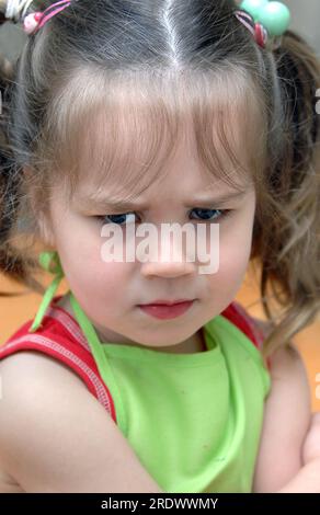 Little girl lets her feelings show with her furrowed brow and crossed arms.  She has pigtails and is wearing a green apron and red shirt. Stock Photo