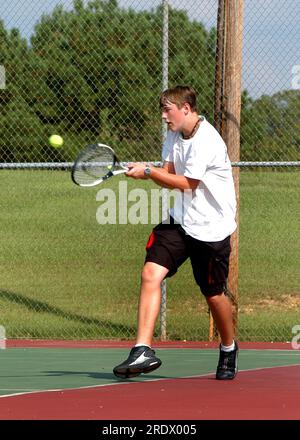 High school varsity tennis player concentrates as he swings his racket to hit the tennis ball.  He is playing on an outdooor court. Stock Photo