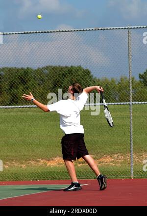 Teenager throws tennis ball high in the air as he serves it.  He is keeping his eyes on the ball and his arm is in the motion of swining to serve. Stock Photo