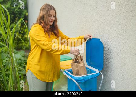 Full size of caucasian woman sorting garbage, throwing a used paper bag in a small recycle bin at outdoor near a home  Stock Photo