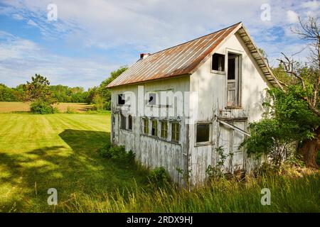 Abandoned farm house in field with busted windows and door next to dead tree Stock Photo