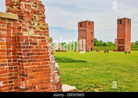 Two freight elevator towers with busted brick pillars and benches in green field with partial wall Stock Photo