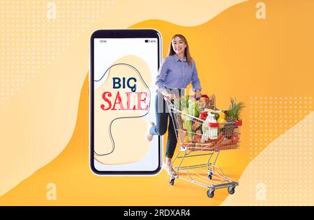 Sale flyer design. Happy woman with shopping cart full of groceries and huge smartphone on color background Stock Photo