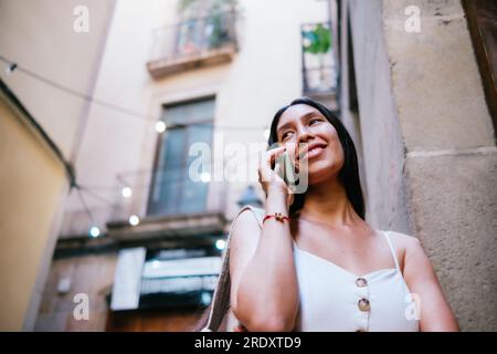 From below glad Latin American female smiling and looking away while leaning on wall of apartment building and answering phone call on street of Barcelona, Spain Stock Photo