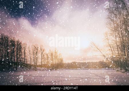 Sunset or sunrise on a frozen pond with birch trees along the banks and falling snow in winter. Snowfall. Vintage camera film aesthetic. Stock Photo