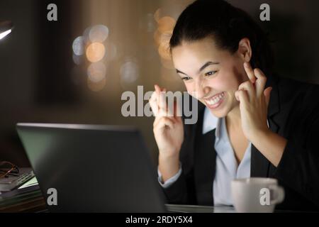 Hopeful businesswoman crossing fingers in the night at office or home Stock Photo