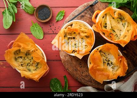 Filo pies with soft feta cheese and spinach in ceramic molds on old red wooden table backgrounds. Filo portions pies. Small Baked Spanakopita pies. To Stock Photo