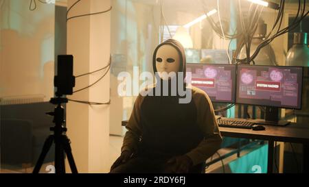 Male spy with anonoymous mask filming live threat video, asking for ransomware instead of leaking important data. Young person threatening to expose information, online cybercrime. Stock Photo