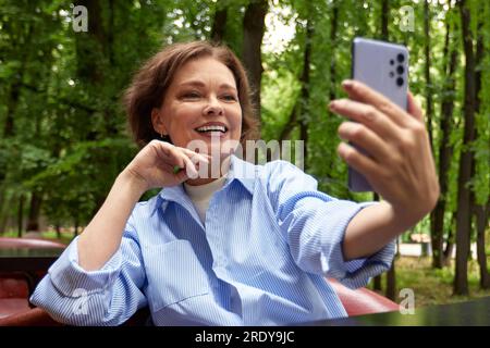 Attractive smiling senior woman making selfie. Mature caucasian lady uses smartphone. Middle aged brunette female in blue shirt enjoying life outside Stock Photo