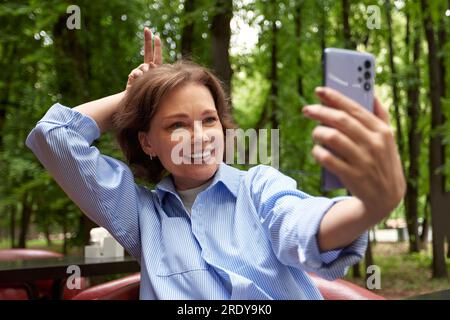 Attractive smiling senior woman making selfie. Mature caucasian lady uses smartphone. Middle aged brunette female in blue shirt enjoying life outside Stock Photo