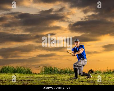 Man in sports clothing hitting baseball with bat in dusk Stock Photo