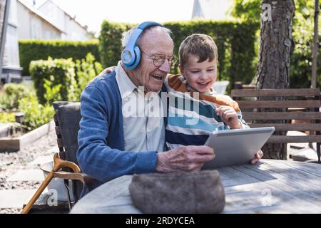 Happy senior man wearing wireless headphones using tablet PC with grandson Stock Photo