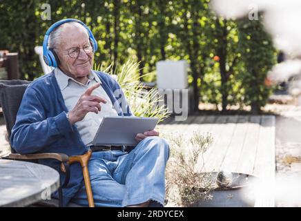 Thoughtful senior man wearing wireless headphones sitting with tablet PC Stock Photo