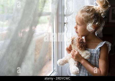 Contemplative girl holding soft toy and looking out of window at home Stock Photo