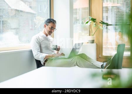 Confident businessman holding coffee cup using laptop working at office Stock Photo