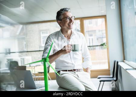 Thoughtful happy businessman holding coffee cup sitting on desk by wind turbine model in office Stock Photo