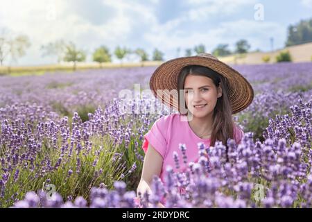 Closeup portrait of beautiful brunette smiling girl in straw hat and pink dress posing in purple lavender field.  Horizontally. Stock Photo