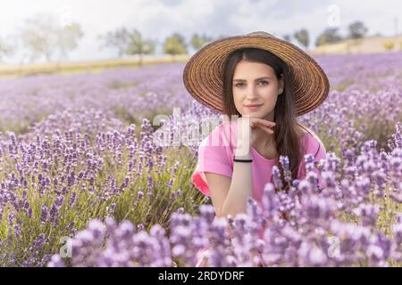 Portrait of beautiful tanned girl in straw hat posing supports her chin in purple lavender field.  Horizontally. Stock Photo