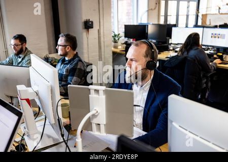 Mature businessman wearing headset working on computer with colleagues at desk Stock Photo