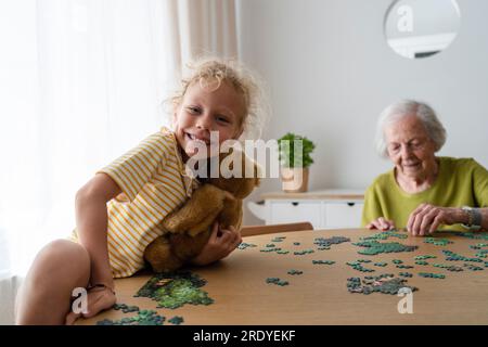 Grandmother solving jigsaw puzzle and granddaughter holding teddy bear Stock Photo
