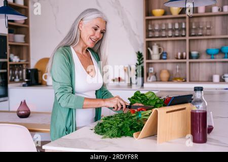 Smiling mature woman watching tutorial and preparing food at home Stock Photo