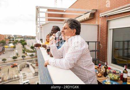Smiling senior man drinking wine leaning on wall with friends in balcony Stock Photo