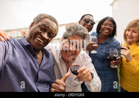 Happy senior man taking selfie with friends enjoying drinks in balcony Stock Photo