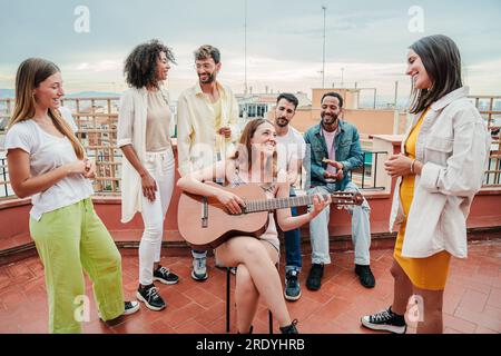 Young adult musician woman playing a guitar and singing a song to her best friends, enjoying a rooftop party with her buddies in the rooftop. Festive meeting of multiracial people having fun together. High quality photo Stock Photo