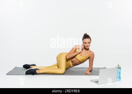 woman talking on smartphone, sitting near laptop and sports bottle on fitness mat, white background Stock Photo