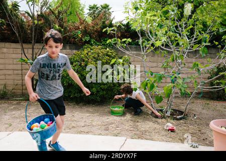 Girl reaches for easter egg from under a tree while boy runs past Stock Photo
