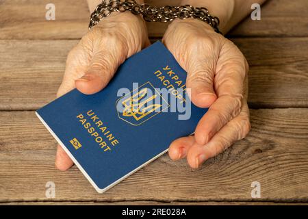 The biometric passport of Ukraine is held in the hands of an elderly woman with her hands tied with a chain on a wooden board, restrictions for a Ukra Stock Photo