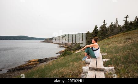 Woman sitting on wooden pathway looking at ocean on a hike. Stock Photo