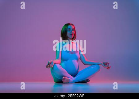 Pregnant woman sitting in lotus pose and doing meditation in neon Stock Photo