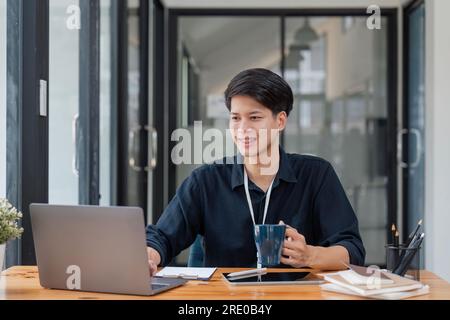 Smiling business man enjoying coffee while working. Young entrepreneur is using laptop. He is sitting at desk in corporate office Stock Photo