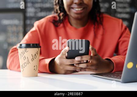 Close-up of African American teacher using smartphone while sitting at table with coffee drink and laptop in the classroom Stock Photo