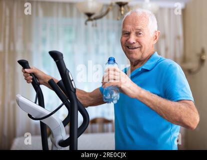 Senior man with bottle of water using elliptical trainer at home Stock Photo