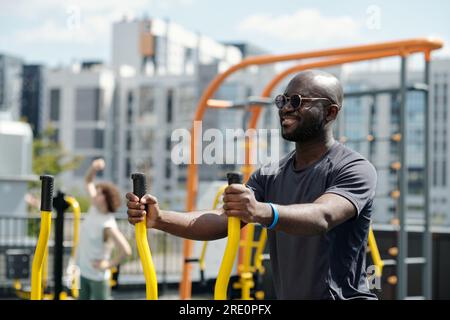 Happy young sportsman in sunglasses and activewear exercising on sports facilities against active woman and urban environment with buildings Stock Photo