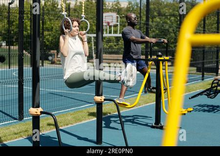 Young brunette sportswoman hanging on still rings and doing L sits on sports ground against African American man exercising on facilities Stock Photo