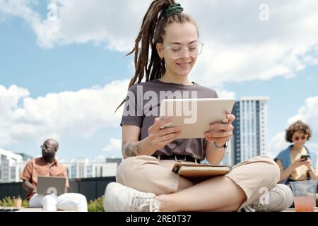 Happy young female student or employee looking at tablet screen while watching online video or working over new project in open air Stock Photo
