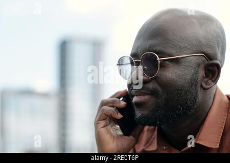 Close-up of young confident African American businessman in eyeglasses holding smartphone by ear while talking to someone outdoor Stock Photo