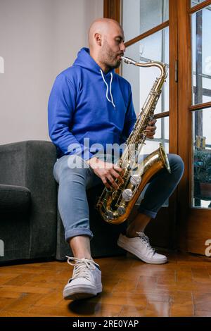 vertical portrait of young venezuelan man in blue playing saxophone sitting on the sofa at home Stock Photo