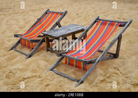 Two empty colorful canvas lounge sling chairs and wooden table on sand beach of resort in Thailand, front side view, high angle Stock Photo
