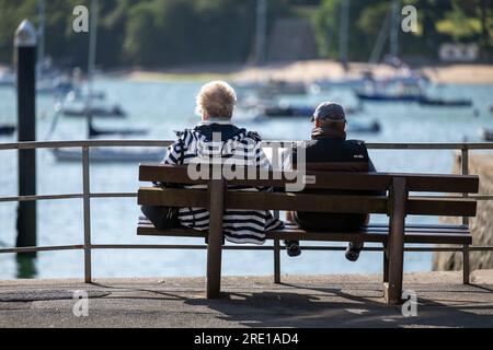 Couple sitting on a public bench in Whitestrand car park, in the centre of Salcombe, Devon, overlooking moored boats in the harbour on a sunny day Stock Photo