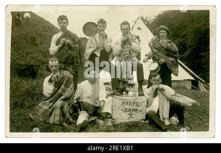 Original early 1920's era postcard of group of good looking young men, messing about whilst camping dressed as Scottish pipers and bandsmen. One of them holds up a greasy frying pan with a charred sausage stuck to it. The lads are wearing tartan - one has a sporran made from a  hair brush attached to the front of his kilt! They have a sign outside their tent stating 'Magpie camp' , unknow location in U.K. possibly Scotland. Stock Photo